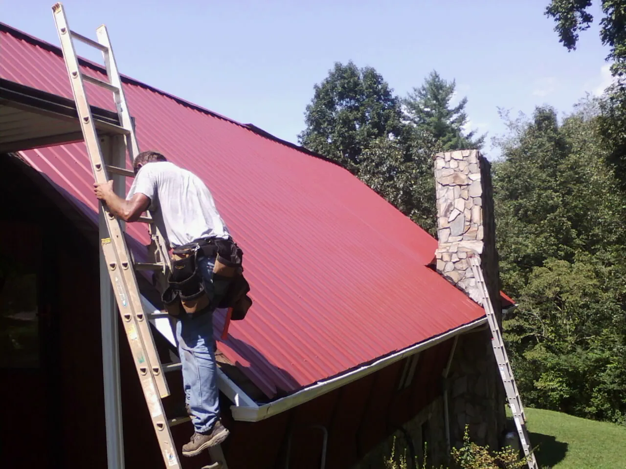 Roofer on a ladder beside a red metal roof and stone chimney — Soffit & Fascia Repair by Roofing Brothers in Westminster
